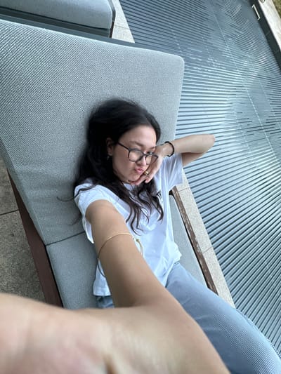 An image taken from a first-person perspective captures a single individual, appearing female, lying on an outdoor lounge chair within Vandœuvres, Switzerland. The subject has dark, wavy hair and wears eyeglasses with dark, possibly tortoiseshell, frames. She is dressed in a white short-sleeved t-shirt and light blue and white striped pants. Her left hand is resting near her chin, and she exhibits a pursed-lip expression while looking directly at the camera. Her right arm, holding the camera, extends into the foreground, partially out of focus, with a thin gold-colored bangle bracelet visible on her wrist. A black beaded bracelet is on her left wrist. She is positioned on a grey upholstered lounge chair with dark wooden or metal frame elements. To the right and upper background, a large, dark grey, slatted surface, consistent with a pool cover or modern outdoor decking, is visible. Light-colored stone or concrete paving is present to the immediate left of the lounge chair. The scene is illuminated by natural light, suggesting daytime.