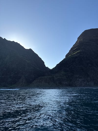 The scene depicts a coastal landscape within the city of Unknown, United States of America. No individuals are present. The lower half of the image is dominated by dark, undulating ocean water, exhibiting rippled reflections of light across its surface. This water meets a dark, rocky shoreline at the base of two large, steep mountains. Small white waves are visible where the ocean interacts with the land. The mountain on the left is predominantly dark, with its upper ridge silhouetted against an unseen bright light source, likely the sun, positioned just beyond the frame to the upper left. The mountain on the right also appears dark and steep, with scattered green vegetation visible on its slopes. A valley or gorge recedes between the two mountains. The upper third of the image shows a clear, light blue sky, brightening towards the area of the implied light source.