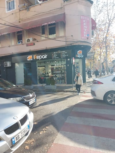 The image captures a sunlit urban street scene in Tiranë, Albania, on what appears to be an autumn day, indicated by the sparse leaves on trees and fallen leaves on the ground. The time of day is likely midday or early afternoon, given the bright light and prominent shadows.
On the left, a multi-story building houses "HOTEL CENTER," prominently displayed on a large sign at the top and again in smaller text on a lower level. Below the main "HOTEL CENTER" sign, a smaller red sign also shows "CENTR..." and part of a phone number, "069...". Various cables are strung across the front of this building.
In the center, a person wearing a light jacket and dark pants stands near the entrance of an "iRepair" store. The store features a large, well-lit "iRepair" sign with a phone icon. Its glass storefront displays numerous electronic items and accessories. Text visible on the window includes "iPHONE," "iWATCH," "iPad," "MacBook," "Servis i shpejte" (fast service), "Garanci" (guarantee), and "Aksesor" (accessories). A list of iPhone models like "X," "XS," "11," "12," "13," "14," and others are also visible.
To the right, a red and white striped crosswalk stretches across the road. Several cars are visible: a white sedan partially on the crosswalk, and another white car's front bumper and grille, bearing an Albanian license plate "AB 228GS," in the foreground. A dark-colored sedan is parked behind it, with a partially visible license plate starting "AA 593...". In the background, other people can be seen walking on the sidewalk.