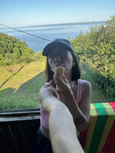 A woman wearing a pink tank top and a gray baseball cap eats an ice cream cone on a balcony overlooking a large body of water and lush green trees. She is holding the cone to her mouth with her right hand, and her left arm is extended toward the camera, likely holding a phone to take a selfie. A colorful striped blanket is draped over the railing of the balcony to her right. The scene is set in Kostovaty, Russia, under a clear blue sky.