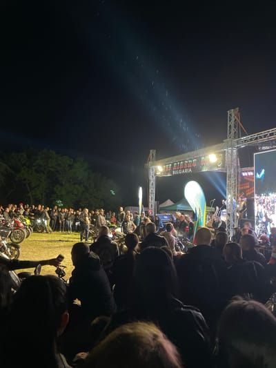 The image depicts a nighttime outdoor gathering, likely a motorcycle rally or event, in or near Chargan, Bulgaria. A crowd of people is gathered around a stage structure with "BULGARIA" emblazoned on the banner. A large screen displays a video feed above the stage. A line of motorcycles is parked on a grassy field near the crowd and the edge of a forested area. The sky is dark.