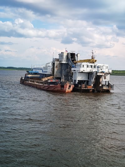 A long, multi-part barge is sailing on a wide, dark river under a partly cloudy sky. The barge is dark brown on its lower hull and white on its upper structures. It is divided into at least two distinct sections connected together. The front section appears to be a long, flat deck with some cargo, including wooden crates, and a person standing near the edge. The rear section is taller and appears to be a working vessel with a crane, a cabin, and various equipment. A Russian flag is visible on the upper part of the rear section. The river has moderate waves, and the banks are lined with green trees in the distance. The overall scene suggests industrial activity or transport on the river, possibly in the city of Krokhino, Russia, known for its hydroelectric power station and river port.