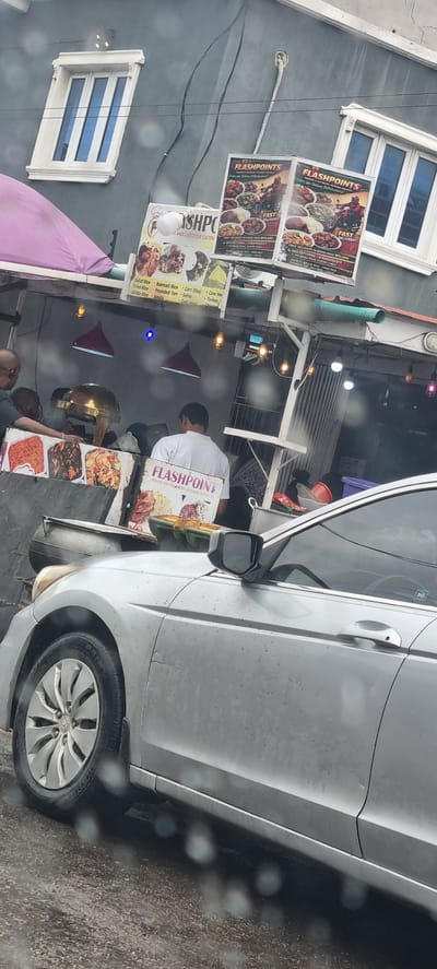 This media file depicts an outdoor scene in Alagbede, Nigeria, likely a food vendor's stall or small eatery. A silver car is parked alongside, partially obscuring the view of the stall. Several signs advertising "FLASHPOINTS INDOOR AND OUTDOOR CATERING SERVICES" are prominently displayed. These signs feature images of various dishes, suggesting a menu of Nigerian cuisine, with text mentioning "Jollof Rice," "Basmati Rice," "Garri (Eba)," "Cow Meat," "Turkey," and "Chicken." One sign also advertises "FAST DELIVERY" with a phone number.

In the background, several people are visible, including a person in a white shirt, likely a vendor or staff member, serving or preparing food. The setting appears to be somewhat cluttered, with food containers and other items. The lighting suggests it is daytime, though the weather appears overcast, with reflections on the car's surface indicating it might be raining or recently rained. The overall impression is a bustling local food business preparing for or serving customers.