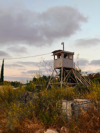 A derelict guard tower stands in a rural area, possibly in the Lachish Regional Council of Israel, surrounded by overgrown vegetation and wild yellow flowers. The tower, made of rusted metal, is elevated on a lattice of supports and features a small cabin at the top with windows. A security camera is mounted on a pole above the cabin. A set of stairs leads up to the cabin, and a power line stretches across the sky in the background. The sky is filled with a mix of light and dark clouds, with a hint of sunset colors. In the foreground, dry grass and rocks are visible. The overall scene evokes a sense of abandonment and decay.