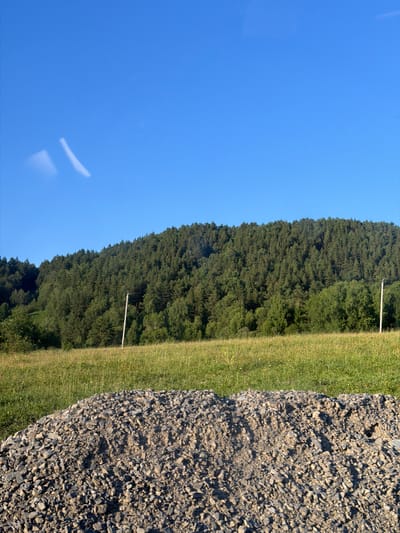 This media file captures a landscape scene in Elekmonar, Russia. The image is oriented vertically. On the left, a vast expanse of clear, bright blue sky dominates the frame. A faint, elongated white streak, possibly a lens flare or a very thin cloud, is visible in the sky.
The right side of the image depicts a transition from a grassy field to a dense forest. The grassy area appears to be a well-maintained lawn, with a strip of mowed grass running parallel to the forest. Two thin, white poles are sparsely visible in the grassy area, suggesting they might be part of a fence or markers.
Beyond the grassy field, a thick, verdant forest covers a hillside. The trees are predominantly dark green, suggesting a coniferous forest. The forest canopy is dense and appears to stretch upwards and outwards.
In the foreground on the far right, there is a close-up view of a rough, gravelly surface composed of numerous small, angular stones of various gray and beige hues. The texture is highly detailed, showcasing individual pebbles and their shadows.
The lighting suggests it is daytime, likely late afternoon or early morning due to the clear sky and the direction of shadows. The weather appears to be clear and sunny. There are no people or animals visible in the image, and no text is present. The overall impression is one of a serene natural environment.