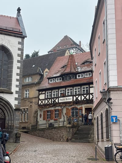 An outdoor street scene in Meissen, Germany, features the Weingut Vincenz Richter building under an overcast sky with traces of snow on rooftops. A cobblestone street slopes downwards in the foreground.
To the left, a large stone building has grey/white ashlar corners, a dark arched entryway, and an arched window above. Its red-tiled roof is topped with a metallic finial.
Center stage is the two-story, half-timbered "Vincenz Richter" building, identified by a sign. Its light grey/beige lower facade has rectangular windows with brown shutters, arched display windows, and decorative dried vines. The upper story displays a darker timber frame with multiple windows. A complex red-tiled roof includes dormers and a prominent conical spire with a green apex. Stone steps, bordered by a rough retaining wall and metal fence, lead to the entrance, where two individuals stand: one in a dark jacket, one in a light grey hooded jacket.
To the right, a multi-story, light pink building with white corner accents has narrow, dark-framed windows. An old-style black street lamp with a security camera is mounted on its corner, with a blue traffic sign (white horizontal bar) below. A trash receptacle and partial bicycle wheel are in the bottom right.
In the background, a light brown building with a dark tiled roof and a circular wrought-iron sign is visible. Further back, another half-timbered building with a brown-tiled gable roof appears. On the far left foreground, two partially obscured individuals are present: one seen from the back with a dark jacket and blue backpack, and another with a visible shoulder and a red object.