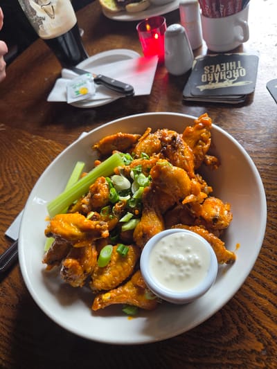 The image shows a white bowl filled with buffalo wings, accompanied by a side of white sauce and celery sticks. The wings are topped with chopped green onions. The bowl sits on a dark wooden table surface. To the left of the bowl, there are condiments (salt, pepper, ketchup) and a stack of coasters with "Orchard Thieves" written on them. Further left, there's a small white plate with a black-handled knife, a lemon or citrus packet, and a glass of Guinness. A small portion of a plate with food on it is also visible in the left corner. An arm and hand are visible in the lower left corner of the frame. The location is suggested to be in Dublin, Ireland.
