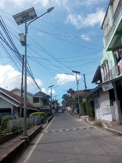 This image presents a street view in Naung Bo Oe, Myanmar, on a clear day with a blue sky and scattered white clouds. The asphalt road, marked with a yellow and black speed bump in the mid-ground, ascends gradually uphill. On the left, a tall grey metal utility pole supports a solar panel array, a street light, and a dense tangle of black electrical wires that crisscross the scene. Adjacent to the road on the left are multiple residential-style buildings, the closest having a tiled roof and white walls, with a black vehicle partially visible parked in front. A low wooden slat fence defines the property line. On the right side of the road, a two-story building with a pale green facade displays clothing hanging to dry from an upper balcony. Below it, a single-story structure with white, woven-textured walls and a visible sign is present. Further up the street, a white car and several indistinct figures are visible, moving or stationary. Additional solar-powered streetlights line the road, and residential buildings, including one with a distinctive tiered roof in the distance, are visible amidst green foliage.