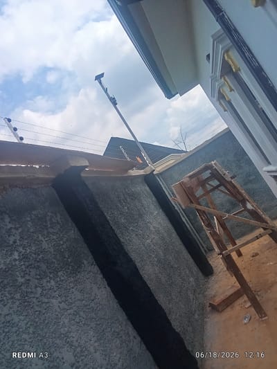 The image depicts an outdoor construction scene. A textured grey wall runs diagonally from the lower left to the upper right of the frame. A dark, vertical gap is visible in the wall. Above the wall, there is an electric fence. In the background, a dark-tiled roof is visible with white trim. A wooden scaffolding structure is positioned to the right of the wall, on sandy ground. A portion of a white building with decorative trim is visible on the far right. The sky is cloudy and blue. The timestamp "06/18/2026 12:16" is visible in the bottom right corner, and "REDMI A3" is in the bottom left. There are no people visible in the image. The location is identified as Nwofia, Nigeria.