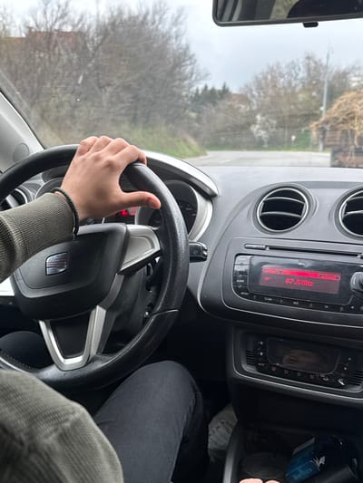 The image depicts a first-person perspective from the driver's seat of a left-hand drive vehicle, in what appears to be a semi-rural or peripheral area within Kumanovo, North Macedonia, under an overcast sky.

A person's right hand, adorned with a dark braided bracelet on the wrist, grips the upper-right section of a black, three-spoke steering wheel featuring a silver SEAT emblem. The driver's arm is covered by an olive-green ribbed sleeve, and their left leg, clad in dark trousers, is partially visible.

The vehicle's dashboard is visible, showing a car radio display tuned to "87.5 MHz" on FM, with "memory FM" and "AF" indicators. Above the radio are two circular air vents. Below the radio, a climate control panel is set to "AUTO." In a storage compartment to the right of the driver, a blue pack of Marlboro cigarettes and a key are present.

Through the windshield, a paved road is visible, curving gently to the left. The roadside is lined with deciduous trees and some green foliage. In the mid-ground on the right side of the road, a vehicle, likely a tractor or utility truck, is pulling a trailer heavily loaded with hay or straw. A small red and white road sign is partially visible on the left side of the road. The windshield shows minor dust or smudges.