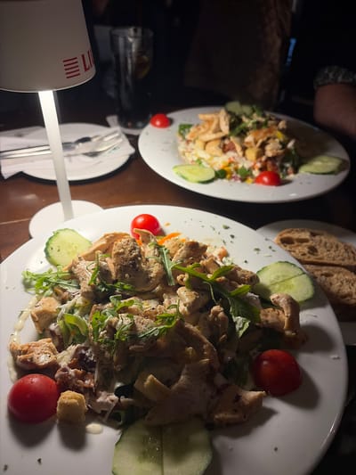 The image depicts an indoor dining scene, likely in a restaurant, featuring two plates of salads with grilled chicken on a dark wooden table. In the foreground, a white, round plate holds a substantial portion of salad composed of cubed grilled chicken, green leafy vegetables (including romaine lettuce and arugula), small red cherry tomatoes, sliced cucumbers, croutons, and a creamy white dressing, possibly with grated cheese. A small, white, modern table lamp with a stylized red "LI" or "LUM" logo and horizontal stripes on its shade illuminates this foreground plate from the left. To the left of the plate, a white paper napkin with a metallic fork rests on the table.
In the background, another white, round plate holds a similar salad with grilled chicken, vegetables, tomatoes, and cucumbers, slightly out of focus. Behind this plate, a tall, clear glass containing a dark beverage, ice, and a citrus slice (lemon or lime) is visible. To the far right, a plate with several slices of light brown bread is present. A person's arm is faintly visible on the right side of the frame, suggesting at least two diners are present. The overall lighting is dim, indicating a nighttime or low-light ambiance, with the table lamp providing focused illumination.
The specific location within the city of Essen, Germany, cannot be determined from the visual information provided in the image.