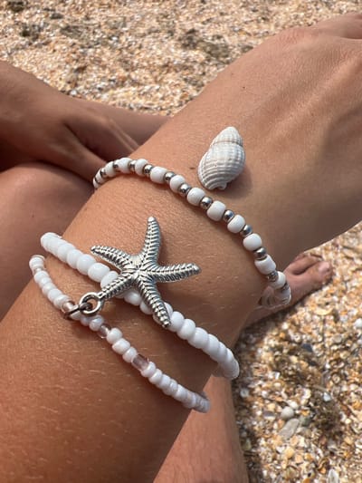 The image shows a close-up of a person's arm with two white beaded bracelets. One bracelet features a silver starfish charm, and the other has a small white seashell. The arm is resting on a sandy surface, likely a beach, with small pebbles and shells scattered around. The lighting suggests it is daytime, with sunlight casting shadows. The background is blurred but shows more of the beach environment, including what appears to be a person's foot in the upper right corner. The overall scene evokes a relaxed, summery, or vacation-like atmosphere. There is no visible text in the image.