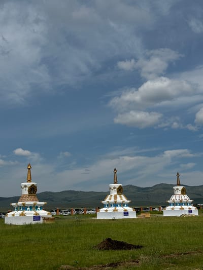 Three white stupas with gold spires stand in a grassy field under a blue sky with scattered clouds. The stupas are ornate, featuring decorative bands of blue and gold, and small figures. In the background, a line of parked white vehicles is visible behind a fence, with rolling green hills beyond. The ground in the foreground is covered in tall green grass, with a pile of dark soil to the right. This scene is located in Orongoi, Russia.