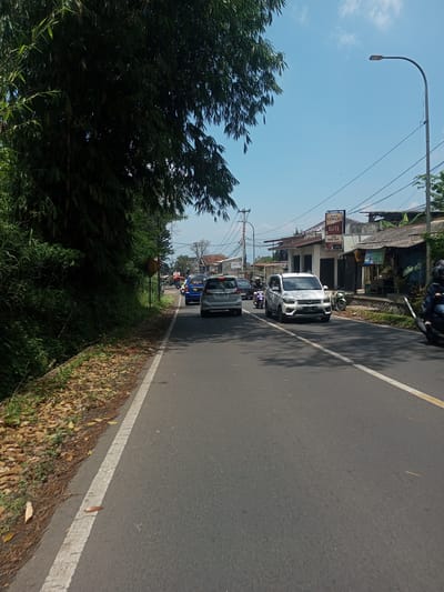 The image depicts a street scene, possibly in a suburban or rural area. Cars are driving on a paved road with a painted dividing line. On the left side of the road, there is dense foliage. On the right side, there are buildings, a street lamp, and overhead power lines. A business sign, partly obscured, is visible, reading "DARLIM COKLAT ELITE TOKO MANG ENOR." A person on a motorbike is on the right side of the road. The sky is blue with scattered clouds.
It is not possible to determine if this image is taken in Nawng Hkar, Myanmar, based on the provided image alone.