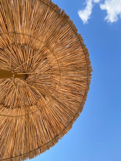 The image shows the underside of a thatched umbrella, made of dried reeds, against a bright blue sky with scattered white clouds. The umbrella is constructed with a central pole and radiating metal supports, with the reeds bound together to form a circular canopy. The texture of the reeds is rough and varied in color, ranging from light tan to a deeper brown. The umbrella occupies the left and lower portion of the frame, with the sky filling the remaining space. There is no indication of a specific location in Balykchy, Kyrgyzstan, nor are there any people or actions depicted.