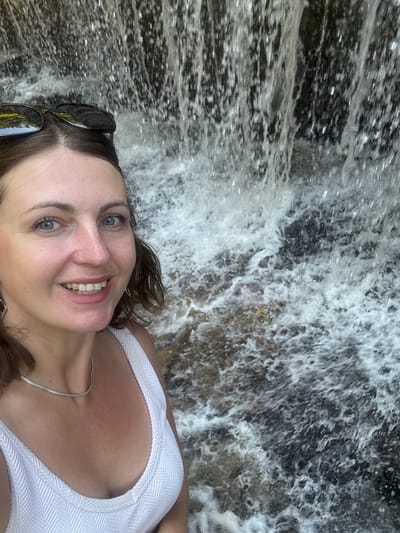 A woman with brown hair and blue eyes smiles at the camera. She is wearing sunglasses on her head and a white top. She is standing in front of a waterfall. The location is not specified in the city of Saint-Claude, France.