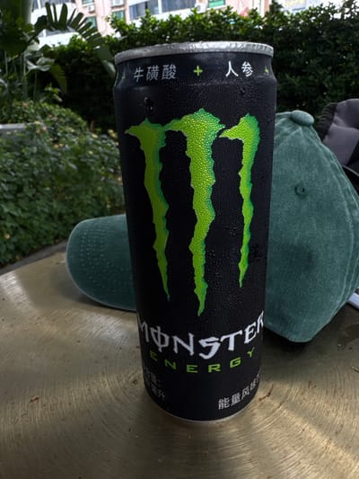 This media file features a Monster Energy drink can with condensation droplets, placed on a textured, metallic surface. The can is predominantly black with the iconic green Monster Energy logo. To the left of the can, partially visible Chinese text reads "牛磺酸" (niú huáng suān), which translates to "Taurine." Further to the right, more Chinese text is visible, reading "能量饮料" (néng liàng yǐn liào), meaning "Energy Drink." Behind the can, a green baseball cap is visible, with a portion of a grey garment peeking out from behind the cap. The background consists of lush green foliage and trees, suggesting an outdoor setting. In the far background, the side of a building with windows can be seen, indicating an urban or suburban environment. The lighting suggests it is daytime. The overall scene is static, with no discernible activity taking place beyond the placement of the objects. The location context provided is Xiangzhou, China.
