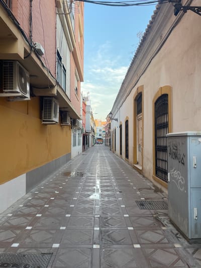 This image shows a narrow street in La Línea de la Concepción, Spain. The street is paved with square tiles featuring a geometric pattern. There are buildings on both sides of the street. The building on the left is painted yellow and has several air conditioning units mounted on its exterior. The building on the right is painted a light beige color and has arched windows with decorative iron bars. A light blue electrical box with graffiti is present on the right side of the street. The street appears to be wet, with puddles reflecting the sky, suggesting recent rain or cleaning. The sky is partly cloudy. In the distance, the street continues, with more buildings and a few people walking.