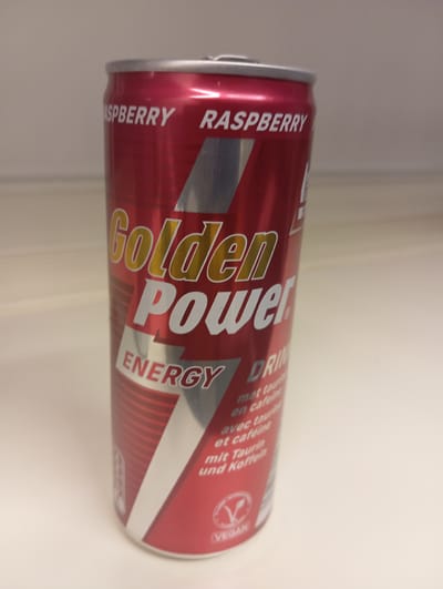 A close-up, slightly angled, eye-level shot captures a single, unopened can of "Golden Power" energy drink. The can is predominantly red with silver accents and text. The brand name "Golden Power" is prominently displayed in a gold, outlined font. Below it, "ENERGY" is written in a silver, outlined font. The can also features text in smaller font, indicating it is a "DRINK" and mentioning "met taurine en cafeine," "avec taurine et cafeine," and "mit Taurin und Koffein," suggesting it contains taurine and caffeine and is available in multiple languages. A silver, diagonal band runs down the front of the can, and a stylized "V" within a circle, signifying "VEGAN," is visible near the bottom. The can sits on a plain, light-colored surface, possibly a table or shelf. The background is out of focus and appears to be a neutral-colored wall. There are no people or animals present in the image. The lighting is even, highlighting the can without harsh shadows. There is no indication of the specific location within Anderlecht, Belgium, beyond the presence of the product itself.