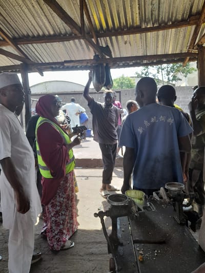 The image depicts an outdoor scene under a corrugated metal roof supported by wooden beams, likely in Kakuri Gwari, Nigeria. Several individuals are present. In the foreground, a person in a white garment and cap is partially visible on the left. To their right, a woman wearing a maroon hijab and a bright yellow high-visibility vest stands, holding a mobile phone and what appears to be a black device. She is looking towards another individual.

In the center of the image, a man in a grey t-shirt and dark pants is holding up a bundle of what appear to be dried fish, suspended from the roof. To his right, a man in a blue t-shirt with Chinese characters on the back is facing away from the camera, looking towards the group. Several other individuals are visible in the background, some in white attire, and one person appears to be holding a blue container.

In the lower right foreground, a weathered mechanical device, possibly a grinder or a press, rests on a dark surface. There are some crumbs or small particles scattered on the surface of this device and the surrounding ground. The environment appears to be a marketplace or an open-air working area, with a dirt or concrete ground and some basic structures. The overall impression is of an active, communal space.