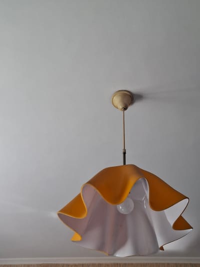 A vintage, yellow and white pendant lamp hangs from a textured white ceiling. The lamp has a distinctive ruffled or petal-like shape, with the outer layer being yellow and the inner layer white. A single, clear lightbulb is visible inside the shade. The ceiling is a plain, off-white color, with a subtle texture. Near the bottom of the image, a horizontal decorative molding or trim is visible along the edge of the ceiling. The scene is indoors, likely in a residential setting. There are no people or animals visible in the image. The lighting appears to be natural, coming from an unseen window or from the lamp itself. The image quality is fair, with some graininess visible, possibly due to low light conditions or the camera used. The exact location within the city of Donetsk, Ukraine, cannot be determined from the visual information provided in the image.