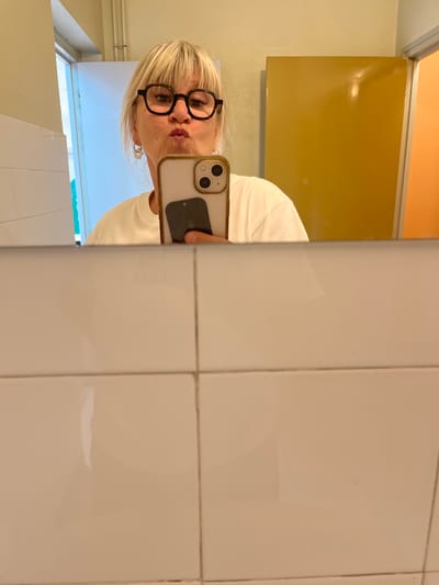 A blonde woman with bangs and glasses is reflected in a mirror. She is wearing a white shirt and has her lips pursed. She is holding up a smartphone to take a selfie. The background includes white tiled walls and doors in yellow and orange. The reflection of a white door is visible on the left side. The scene appears to be a restroom in a public facility located in La Fouillouse, France.