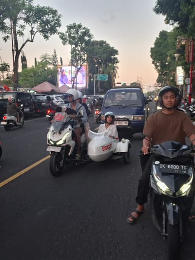 The image shows a street scene in Gianyar, Indonesia, likely during dusk. A man wearing a brown shirt and a black helmet is riding a black motorcycle with a license plate reading "DK 6000 TG". Beside him, a white motorcycle with a sidecar is being ridden by a man in a blue shirt and white helmet, carrying a child in the sidecar who is wearing a white shirt and blue helmet. The sidecar has "BALI SIDE CAR" written on it. In the background, various vehicles, including cars and other motorcycles, are present in traffic. Trees line the street, and a large digital billboard displaying people is visible in the distance. The sky has a hazy, twilight appearance.