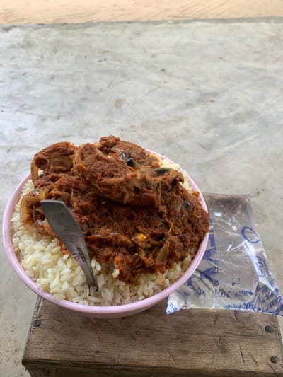 The scene depicts a meal set on a wooden surface in Ikot Ete Ekpo, Nigeria. A light pink-rimmed bowl, positioned centrally, contains white rice topped with a generous portion of reddish-brown stew. The stew features visible pieces of meat or fish and sliced onions. A silver-colored metal spoon is vertically inserted into the food within the bowl. To the right of the bowl, a clear plastic sachet of drinking water, partially filled, rests on the same wooden surface. Blue markings and a droplet logo are visible on the sachet. The wooden surface itself is light brown, appears aged, and is composed of at least two planks, secured by visible nail heads. The background consists of a light grey, textured ground surface, possibly concrete or packed earth, illuminated by natural light. No individuals are present in the image.
