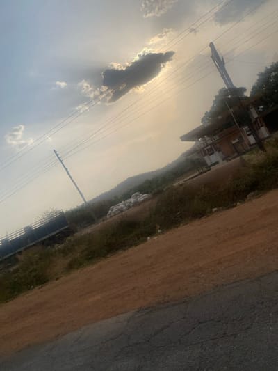 A tilted shot captures a rural scene under a bright, hazy sky. Power lines crisscross the sky, partially obscuring a cloud formation resembling a figure. A building sits atop a hill, framed by trees and greenery. A dirt road runs parallel to a paved road in the foreground. The overall color palette is dominated by shades of brown, green, and sky blue.
