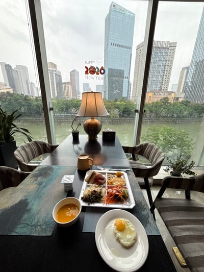 The image captures an interior dining scene overlooking a river and a bustling cityscape, specifically in Jinjiang District, Chengdu, China. The vantage point is from a room with expansive, floor-to-ceiling windows that provide a panoramic view.
In the foreground, a dark, polished dining table is set for breakfast. On the table, to the left, is a textured placemat in shades of blue and green. A white bowl contains an orange-colored soup or puree, and next to it is a small plastic cup of yogurt. A white, rectangular serving tray holds various breakfast items: triangular sandwich halves, slices of processed meat, a portion of rice mixed with green peas, a small salad with shredded carrots, and a small round, fried item. A fork and knife are visible near the tray. To the right, a separate white plate presents a perfectly cooked sunny-side-up fried egg. Further back on the table, a yellow mug contains a beverage, and a decorative table lamp with a cream-colored shade and an ornate bronze-toned base provides warm light. A tissue box with a leopard print pattern is situated behind the lamp, and a small potted plant with green leaves sits near the window. Four chairs with striped gray and white fabric upholstery are arranged around the table, two of which are clearly visible.
Through the large windows, the outside environment is visible under an overcast sky. A wide, green-tinged river, consistent with the Jinjiang River, flows directly below, lined with dense green trees and vegetation on both banks. Beyond the river, a dense urban landscape extends with numerous high-rise buildings of varying architectural styles and heights. Prominent among them is a very tall, light blue glass skyscraper with vertical dark window strips and a distinctive pointed or tiered top, likely a landmark in Chengdu's financial district. Another building on the left has "四川省" (Sichuan Province) characters visible on its facade. A "HAPPY 2026 New Year" decoration, featuring stylized numbers with animal motifs (the '6' resembling an auspicious Chinese animal), is affixed to the window pane. No people are visible in the scene. The overall impression is one of a serene breakfast setting within a vibrant urban environment.
