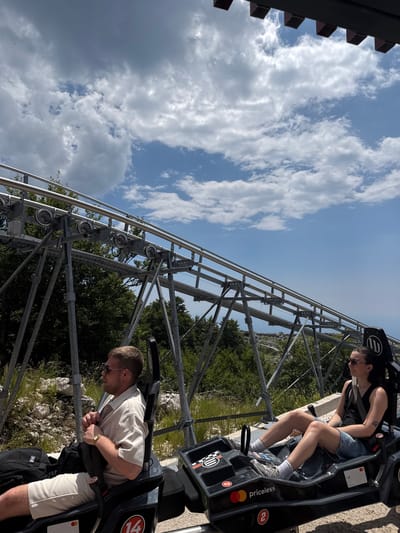 This media file depicts two individuals on a roller coaster or similar gravity-powered ride, likely at an outdoor attraction. The perspective is from a slightly elevated angle looking down at the riders and the track. The sky is a bright blue with scattered white clouds, suggesting it is daytime and the weather is fair. Lush green trees and rocky terrain are visible in the background, hinting at a natural or mountainous location.
The person in the foreground, a man, is wearing a light-colored collared shirt and beige shorts. He is seated in the ride's cart, leaning forward, and appears to be gripping the safety harness. His expression is difficult to discern, but his posture suggests anticipation or excitement. He is wearing sunglasses and a wristwatch.
In the background, another individual, a woman, is seated in a cart ahead of the man. She is wearing a black tank top, denim shorts, and sunglasses. Her head is turned slightly, and she also appears to be looking forward.
The ride's structure is prominently featured, with metal tracks and supports visible. The carts are dark-colored with some branding. Text visible on the carts includes "priceless," a Mastercard logo, and the number "2" within a red circle. Another cart shows the number "14" in a similar red circle. The overall scene conveys an atmosphere of outdoor recreation and thrill-seeking. The location context of Žanjev Do, Montenegro, suggests this might be a specific tourist attraction in that region.