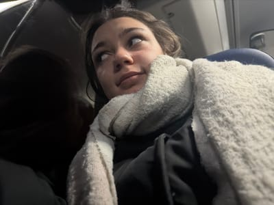 A person wearing a thick, textured scarf is looking directly at the camera. They appear to be seated, with part of an airplane seat visible. Their eyes are gazing upwards. The lighting is somewhat dim, casting shadows on the upper portion of the image. The individual has fair skin and brown hair.