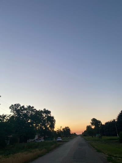 The image depicts a rural road in Repyakhovka, Russia, during twilight. The sky transitions from a pale blue at the top to a warm orange hue near the horizon, indicating sunset. On the left side of the road, there are several houses partially obscured by dense trees and overgrown vegetation. A white car is parked in front of one of the houses. Further down the road, two distant figures can be seen walking away from the camera. On the right side of the road, there are more trees and power poles. The road itself appears to be asphalt with some visible cracks and patches. The overall atmosphere is serene and quiet, characteristic of a rural evening.