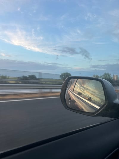 The image presents a serene landscape captured from inside a moving vehicle, likely on a highway near Óföldeák, Hungary. The time of day appears to be either sunrise or sunset, indicated by the warm, soft light and the hues in the sky.
On the right side of the frame, the dark window frame and door panel of the car are visible, providing context for the interior perspective. Dominating the mid-right is the car's side mirror, which offers a captivating reflection of the scene behind the vehicle. Within the mirror, the sky glows with vibrant golden, orange, and soft pink tones, interspersed with delicate clouds, highlighting the golden hour. Reflections of trees lining the road and the asphalt surface of the highway are also visible within the mirror.
The broader view outside the car, particularly to the left and top of the frame, showcases a vast blue sky. It is dotted with scattered white, wispy clouds and some larger, softer grey cloud formations. Below the horizon, a section of a highway or major road is visible, featuring a metal guardrail extending into the distance, with trees and fields beyond.
No people are visible in the image, and there is no discernible text. The scene conveys a sense of peaceful travel, observing the beauty of the sky and surroundings during a journey.