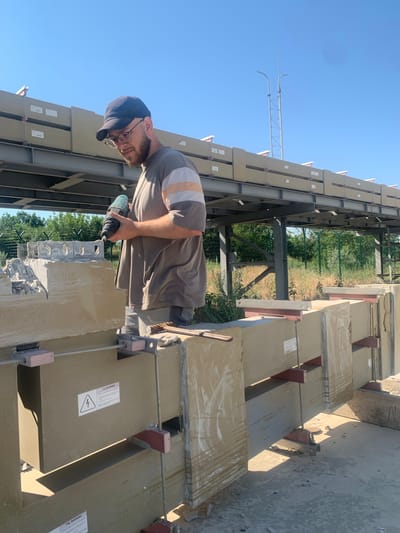 The image shows a construction worker in Pivdennoukrainsk, Ukraine, using a drill on a large, light-colored concrete block. The worker, wearing a dark baseball cap, glasses, and a t-shirt with a striped pattern on the sleeve, is positioned in the foreground. Several similar concrete blocks are arranged in a row, supported by metal and red brackets, with cables running through them. In the background, a multi-level metal structure holds more of these blocks. Beyond the construction area, trees and a fence are visible under a clear blue sky. A metal tower with lighting fixtures stands in the distance. Warning labels, one with an electrical hazard symbol, are affixed to some of the blocks. The ground is concrete and dirt.