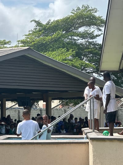 The image depicts an outdoor baptismal service in Itoga, Nigeria. In the foreground, three men in white t-shirts are positioned around a baptismal pool. One man, seen from the back, is partially submerged in the water, with another man on his left and a third man on his right, who is holding a metal railing. Behind them, a crowd of people, dressed in various attire, is gathered, observing the ceremony. The background features a covered structure with a corrugated roof and wooden beams, with lush green trees and a cloudy sky overhead. The overall scene suggests a religious gathering within a community setting.