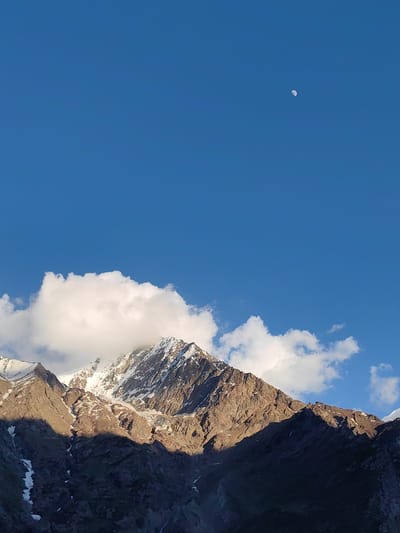 The image depicts a mountainous landscape under a clear blue sky. In the upper right quadrant of the sky, a waxing gibbous moon is visible. The foreground is dominated by rugged, snow-capped mountains. Sunlight illuminates the upper ridges and peaks, casting deep shadows on the lower slopes. Patches of snow are visible on the rocky surfaces, and some clouds drift near the mountain tops. The scene conveys a sense of natural grandeur and is likely a daytime photograph given the bright sunlight. The location is not specified as Kead, India, but rather as a mountainous region in general.