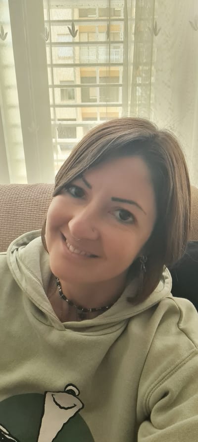 A selfie of a middle-aged woman with short brown hair, wearing a light green hooded sweatshirt with a cartoon graphic, a beaded necklace, and a nose ring. She is smiling. The background includes a window with sheer curtains and a building exterior visible through the window. Based on the architectural style of the buildings and window design, the location is likely Nahariyya, Israel. The woman is indoors.