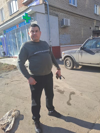 The scene depicts an adult male standing on cracked asphalt, facing the camera, on a clear, sunny day in front of a commercial building in Меткомелино, Russia. The man, appearing middle-aged with short dark hair, wears a dark olive-green long-sleeved sweatshirt with white text, black athletic pants, and dark sneakers with red accents. He has a dark watch on his left wrist. His left hand is in his pocket, while his right hand holds a black smartphone and car keys.
Behind the man to his left is a "ЗДРАВСИТИ АПТЕКА" (Zdravcity Pharmacy) with large glass windows and doors, illuminated by blue neon lighting and topped with a prominent green cross sign. A partially visible street sign above the pharmacy reads "Плеханова 11К1". The building itself is two-storied, constructed of light-colored brick, with white-framed windows. An external air conditioning unit is mounted on the brick wall to the right of the pharmacy entrance.
To the man's right, a dirty, dark-colored SUV, resembling a Lada Niva, is parked on the street. It features a visible roof rack and is covered in dust or mud. On the ground near the man's left foot, a crumpled, light-colored plastic bag is visible. The asphalt surface exhibits multiple cracks and what appear to be wet or oily stains. The strong sunlight casts distinct shadows of the man and the vehicle to the right.
