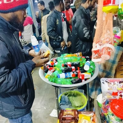 The image captures a lively scene at a street vendor's stall, likely in a market or bustling public area, possibly in South Asia.
In the foreground, a man with a beard, wearing a dark blue jacket and a red and blue knitted cap, is engrossed in consuming food and drink. He holds a piece of yellowish sponge cake or pastry in his right hand and a white plastic bottle with a blue label in his left hand. The bottle appears to be a dairy or yogurt drink, with Bengali text "প্রাণ" (Pran) clearly visible on its label, indicating a Bangladeshi brand.
Central to the image is a large, metallic, round basin brimming with numerous small, single-serving plastic bottles of various beverages. Many of these bottles are clear green, some are dark brown or black (identifiable as Coca-Cola by their labels), and a few are white or blue, some bearing the text "Lacchi" (a popular South Asian yogurt drink). These bottles are piled high, suggesting a wide selection for customers.
Below the main beverage display, the vendor's cart or stand extends downwards, revealing more items. A light green plastic basket holds what appears to be green leafy vegetables or herbs, along with a blue cloth. Further down, there are brown packaged items, possibly biscuits or snacks, with the English text "ALL TIME" visible on one package. Next to these are yellow-packaged goods.
On the far right, a mesh net bag hangs, containing several small red and white disposable cups, possibly for serving drinks. Below this, a clear plastic container is filled with an assortment of colorful candies or small toys, and a red plastic lid or container is also visible.
In the background, several other men are present, some partially obscured. They appear to be patrons or passersby, adding to the bustling atmosphere. One man in a black jacket stands behind the foreground man, while another further back wears glasses. The overall lighting suggests an indoor setting or evening time, relying on artificial illumination. The scene depicts a casual transaction or a brief stop for refreshment in a busy urban environment.