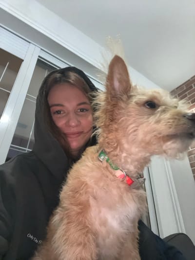 A woman in a hooded sweatshirt smiles at the camera while holding a fluffy, light-brown dog. The dog has a red and green patterned collar. A window with vertical blinds is in the background. Brick is visible at the top corner of the frame. The woman's sweatshirt has "Only Do Story" printed on the lower right side. The image appears to be a selfie taken indoors.