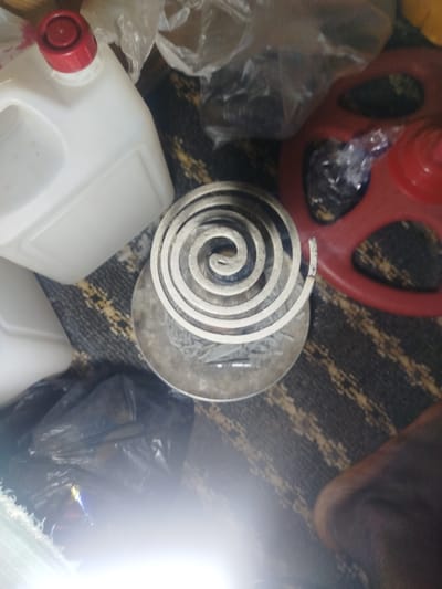 A spiral mosquito coil rests in a circular, metallic dish on a dark, patterned carpet. To the left of the coil is a white plastic jerrycan with a red cap. Behind the jerrycan are various plastic bags and a red plastic object. To the lower left, a black plastic bag partially obscures a bright light source. To the lower right, a dark-skinned limb, possibly an arm or leg, is visible. The scene appears to be indoors.