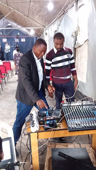 The image shows two men standing in a white tent. The man on the left is wearing a black blazer over a white shirt and blue jeans. He is pointing at an electrical device with a white power outlet attached to a wooden table. The man on the right is wearing a red and white striped sweater and blue jeans. He is standing beside the table, looking down at the device. There is a sound mixing console on the table, which has multiple wires connected to the electrical device. There are multiple electrical wires connecting the device to the other end of the table where a black laptop and a white box are placed. The background shows a number of red chairs and people standing at the far end of the tent. The location is Ota, Nigeria, based on coordinates (latitude: 6.686, longitude: 3.176).