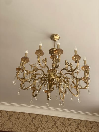 This is a medium shot, angled photograph of a decorative, ornate chandelier mounted on a ceiling. The chandelier is made of a golden-colored metal and features multiple arms adorned with clear and amber-colored crystals. Each arm has a candelabra-style light fixture with a white bulb. The chandelier has a classic, elegant design with swirling metal accents.
The ceiling appears to be a light, neutral color, possibly white or off-white. To the right of the chandelier, a section of wallpaper is visible. This wallpaper has a damask or baroque pattern in shades of brown and gold. A white architectural molding or trim separates the ceiling from the wallpapered wall. The lighting in the image suggests it is indoors, and the overall ambiance is one of luxury and sophistication. No people are visible in the photograph. The image does not contain any visible text. The context provided is Bogatyryovka, Russia, which might indicate an indoor residential or commercial setting within that location.