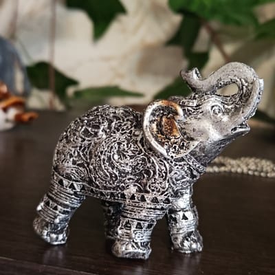 A silver-colored elephant figurine is on a dark wooden surface. The elephant has intricate designs on its body and legs. The trunk is raised, and one ear has some brown discoloration. The background is blurred, showing greenery and other objects out of focus. There is a beaded necklace behind the elephant. The location is unspecified as nothing indicates that this is Novo-Nikol'skoe, Russia.