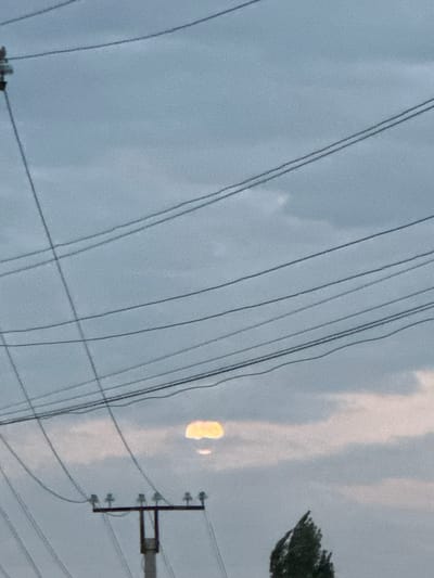 The image depicts a sky filled with numerous power lines crisscrossing the frame. In the distance, a hazy, luminous orb, possibly the moon or a bright light source, is visible through a break in the clouds. In the foreground, a power pole with insulators stands vertically, supporting more power lines. A silhouetted tree is partially visible on the right side of the image, its foliage appearing windswept. The overall impression is a somewhat industrial or utilitarian urban landscape under a cloudy sky.