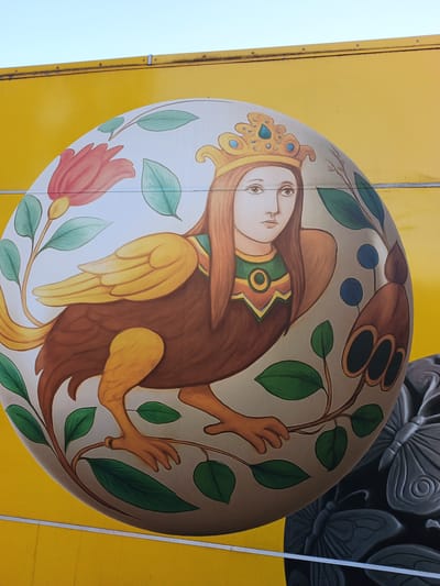 The image shows a mural painted on a yellow wall, likely part of a building. The mural depicts a mythical creature with the torso and head of a human woman and the body of a bird. The woman has long brown hair, wears a golden crown adorned with blue jewels, and has a patterned collar. Her bird body is brown with golden wings and taloned feet. She is surrounded by stylized plants and flowers, including a large pink bloom and green leaves. To the right of the central figure, a section of another mural is visible, featuring detailed grey carvings of butterflies. The mural appears to be located on the exterior of a building in Yaroslavl, Russia, though specific location details are not provided in the image itself. The overall scene is brightly lit, suggesting it is daytime.