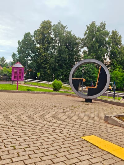 This is a photograph taken in Khotkovo, Russia, featuring a public space with a circular seating structure and a pink public bookshelf. The foreground is paved with light brown brick pavers, with patches of grass growing between them. A bright yellow tactile paving strip is visible on the right side of the foreground. In the middle ground, the circular seating structure, made of dark metal with wooden accents, is prominent. It contains a seat and a small table-like surface within its circular frame. To the left of the seating structure, a pink, house-shaped public bookshelf with glass doors stands on a pole. A ramp with a metal railing is visible behind the bookshelf and seating structure. The background consists of lush green trees and a cloudy sky. Several lampposts are scattered throughout the scene.