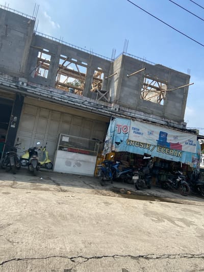 The image shows a street scene in Margaasih, Indonesia (latitude: -6.942, longitude: 107.551). There is a partially constructed building with exposed wooden framing and concrete blocks. In front of the building, several motorcycles are parked. A shop selling groceries and other items is located at the bottom right of the image. The shop sign reads "TOKO" (Indonesian for "store") and "GROSIR / ECERAN" (Indonesian for "wholesale/retail"). The street is paved concrete and there is a small puddle of water in front of the shop. The sky is mostly clear, with a few wispy clouds. A building across the street is visible in the background. No people are visible.