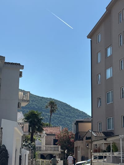 This image captures a view of a town or village nestled against a lush, green mountainside under a clear blue sky. The perspective is unusual, with the buildings appearing to be at the top of the frame, looking down onto the scene. A prominent object in the sky is a white contrail left by an airplane, stretching diagonally across the frame.

Several buildings are visible, constructed in a style that suggests a Mediterranean or coastal location. One large building, with a beige facade, features a grid of numerous rectangular windows. Below this, other buildings with lighter colored facades and balconies can be seen. Some structures have terracotta tile roofs.

Vegetation plays a significant role in the scene, with palm trees scattered throughout. One palm tree stands tall in the middle ground, with its fronds fanned out. The mountainside is densely covered with trees, indicating a natural, possibly somewhat undeveloped, setting.

The time of day appears to be daytime, likely late morning or early afternoon, given the bright sunlight and the clear sky with no visible clouds. The weather seems pleasant and sunny.

There are a few people visible. In the lower right, a person wearing a white top and shorts is standing. Further back, other figures can be seen in the vicinity of the buildings, suggesting a leisurely, everyday atmosphere.

The overall impression is one of a sunny day in a scenic location, possibly a holiday destination, with a blend of urban development and natural beauty. The rotated perspective adds a unique and slightly disorienting element to the composition. No text is visible in the image.